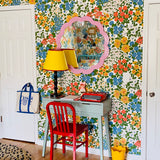 A colorful room corner featuring a retro floral wallpaper wall, a light blue desk, a red chair, a yellow lamp, and a pink scalloped mirror.
