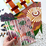 A hand with red nail polish holds a square fabric swatch featuring a pink, green, and orange dotted pattern. In the background, several other patterned fabric swatches are arranged on a white surface, including a geometric orange, yellow, and blue design, a large floral print in brown, orange, and green, and a white pattern with small orange berries.