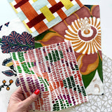 A hand with red nail polish holds a square fabric swatch featuring a pink, green, and orange dotted pattern. In the background, several other patterned fabric swatches are arranged on a white surface, including a geometric orange, yellow, and blue design, a large floral print in brown, orange, and green, and a white pattern with small orange berries.