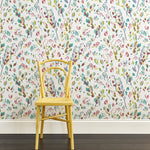 A yellow chair stands in front of a wall papered in a painterly leaf print in purple, blue, green and white.