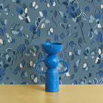 Bright blue vase stands in front of a wall papered in a painterly leaf print in shades of blue.
