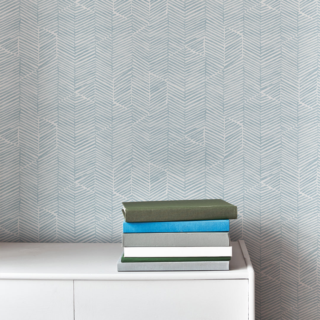 An end table with a stack of books stands in front of a wall papered in a dense herringbone print in light blue and white.