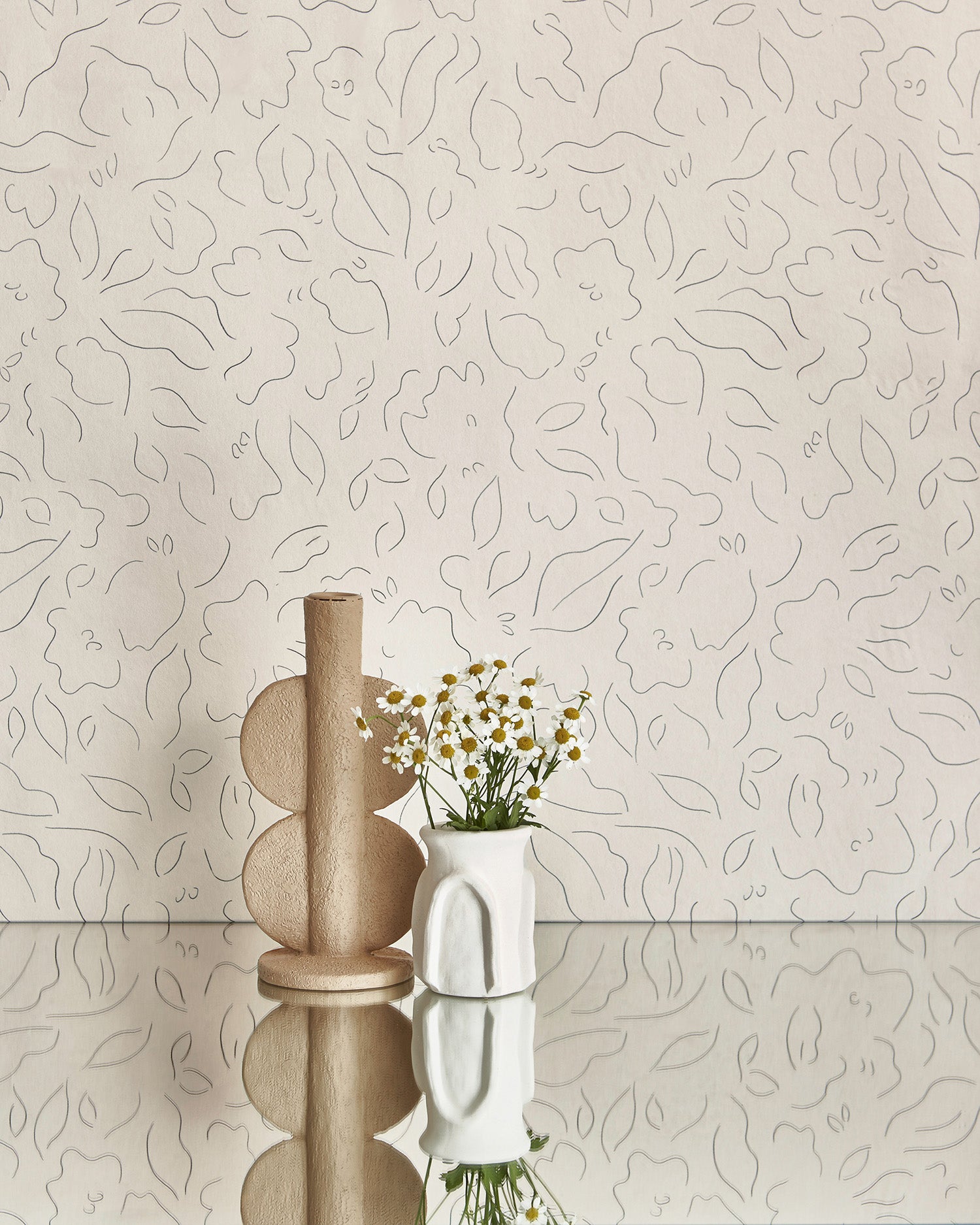 Two vases stand in front of a wall papered in a minimalist floral print in black on a cream field.