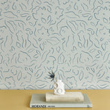 A stack of books and vase stand in front of a wall papered in a minimalist floral print in navy on a turquoise field.