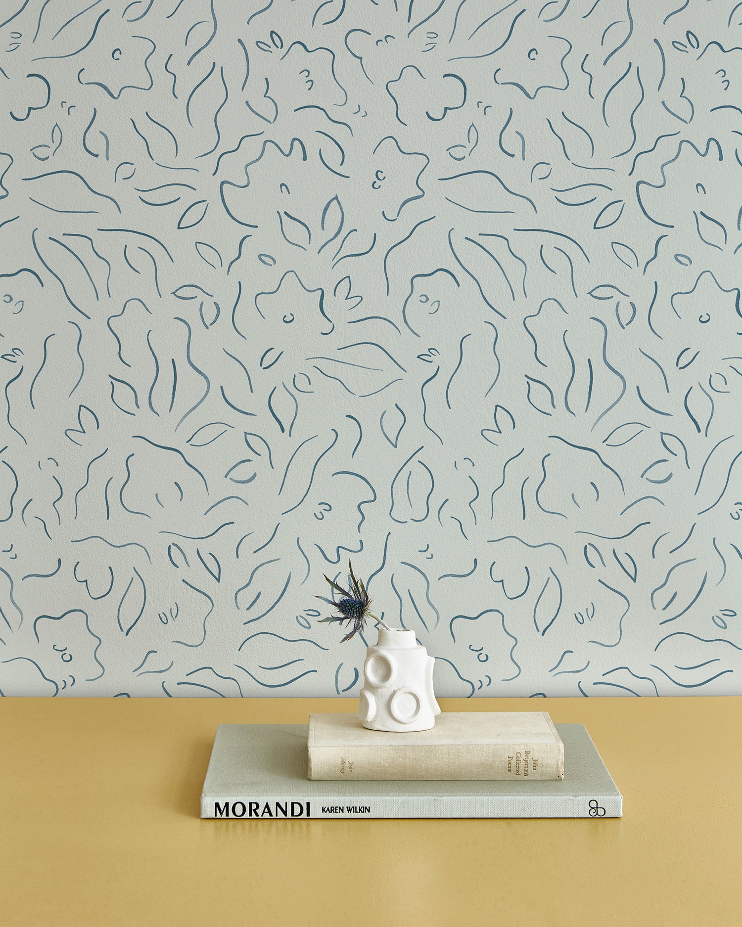 A stack of books and vase stand in front of a wall papered in a minimalist floral print in navy on a turquoise field.