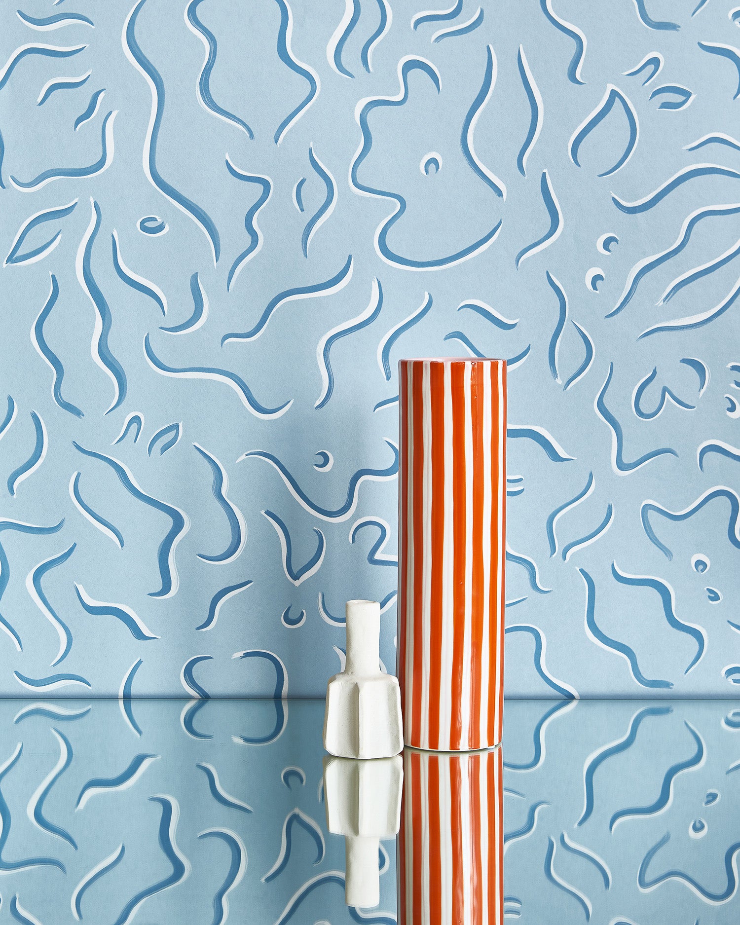 Two vases stand in front of a wall papered in a painterly floral print in blue and white on a light blue field.