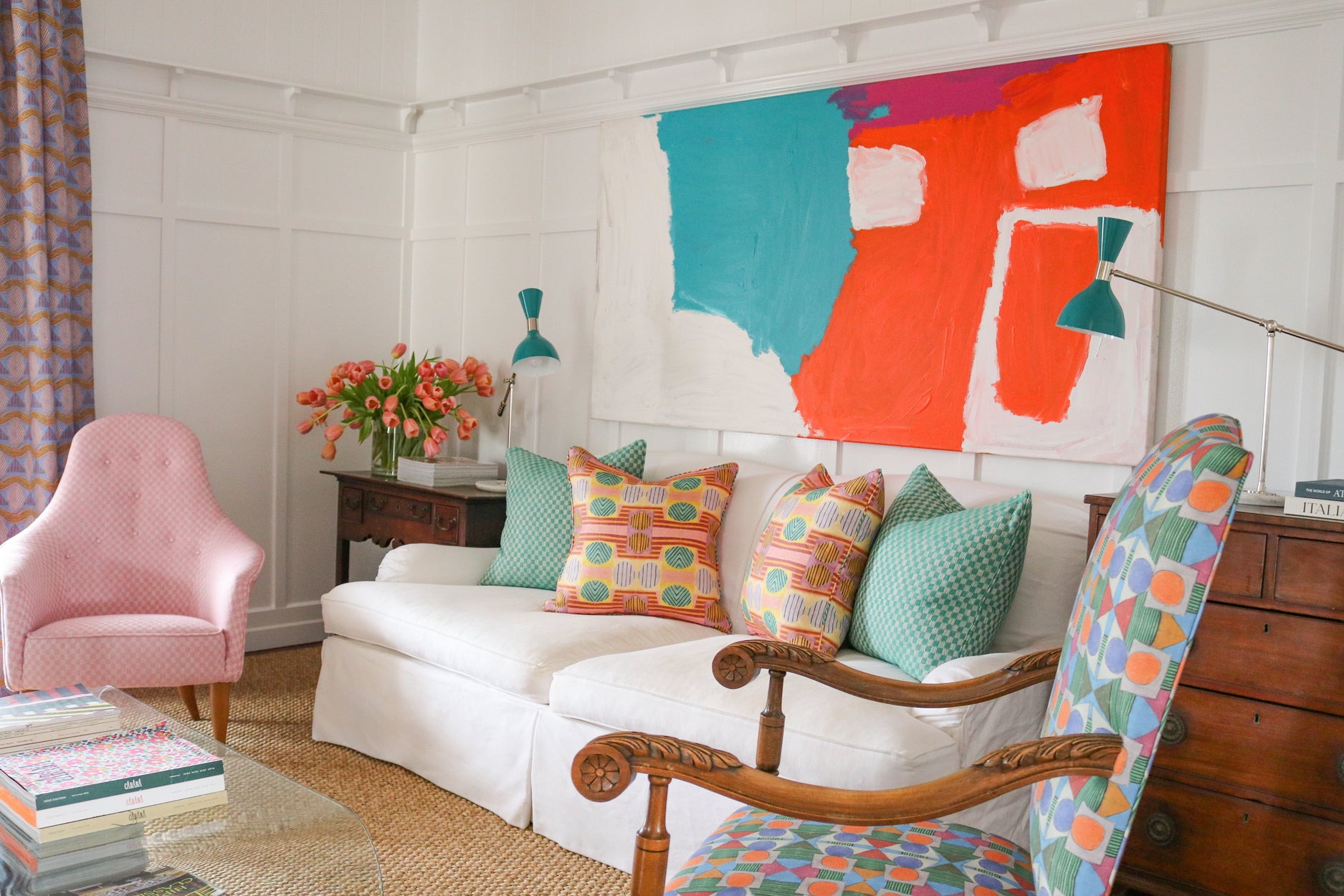 Detail of a living room scene with pink upholstered side chairs, plexiglass coffee table with stacked magazines below and on top with a vase of pink tulips, White couch with bold patterned throw pillows against a white wall with decorative paneling and a large abstract painting in teal, white and red. 