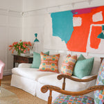 Detail of a living room scene with pink upholstered side chairs, plexiglass coffee table with stacked magazines below and on top with a vase of pink tulips, White couch with bold patterned throw pillows against a white wall with decorative paneling and a large abstract painting in teal, white and red. 