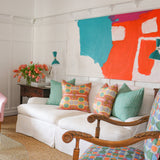 Detail of a living room scene with pink upholstered side chairs, plexiglass coffee table with stacked magazines below and on top with a vase of pink tulips, White couch with bold patterned throw pillows against a white wall with decorative paneling and a large abstract painting in teal, white and red. 
