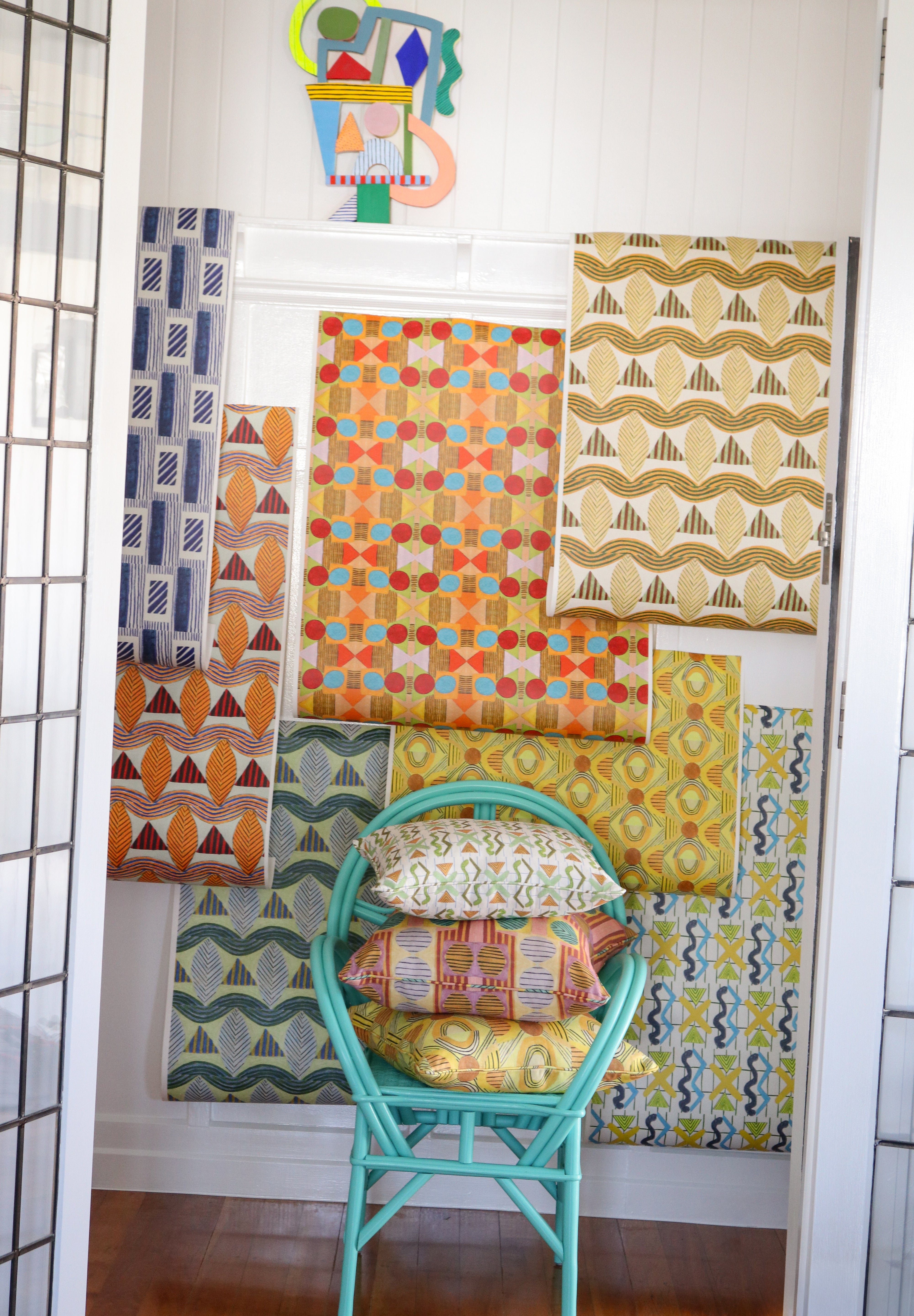 Interior setting looking through a doorway to with a turquoise rattan chair with printed throw pillows against a white wainscoted wall with a collage of geometric patterned wallpapers in bright colors. 