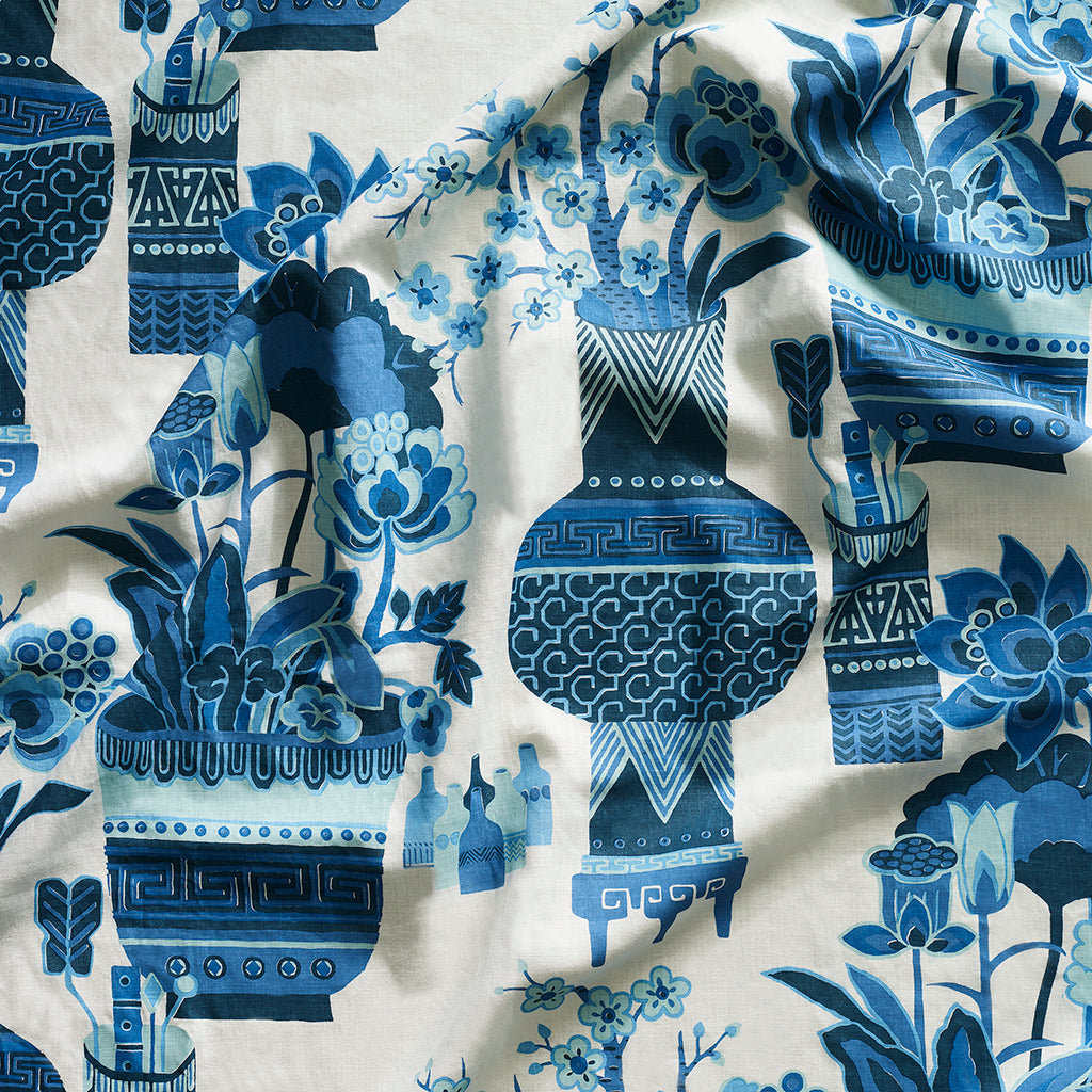 Close up of gently draped fabric in an ornate hand drawn urn and floral pattern in shades of blue on white. 