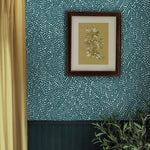 A floral painting hangs on a wall papered in a scrolling stem and leaf print in turquoise on a white field.