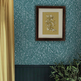 A floral painting hangs on a wall papered in a scrolling stem and leaf print in turquoise on a white field.