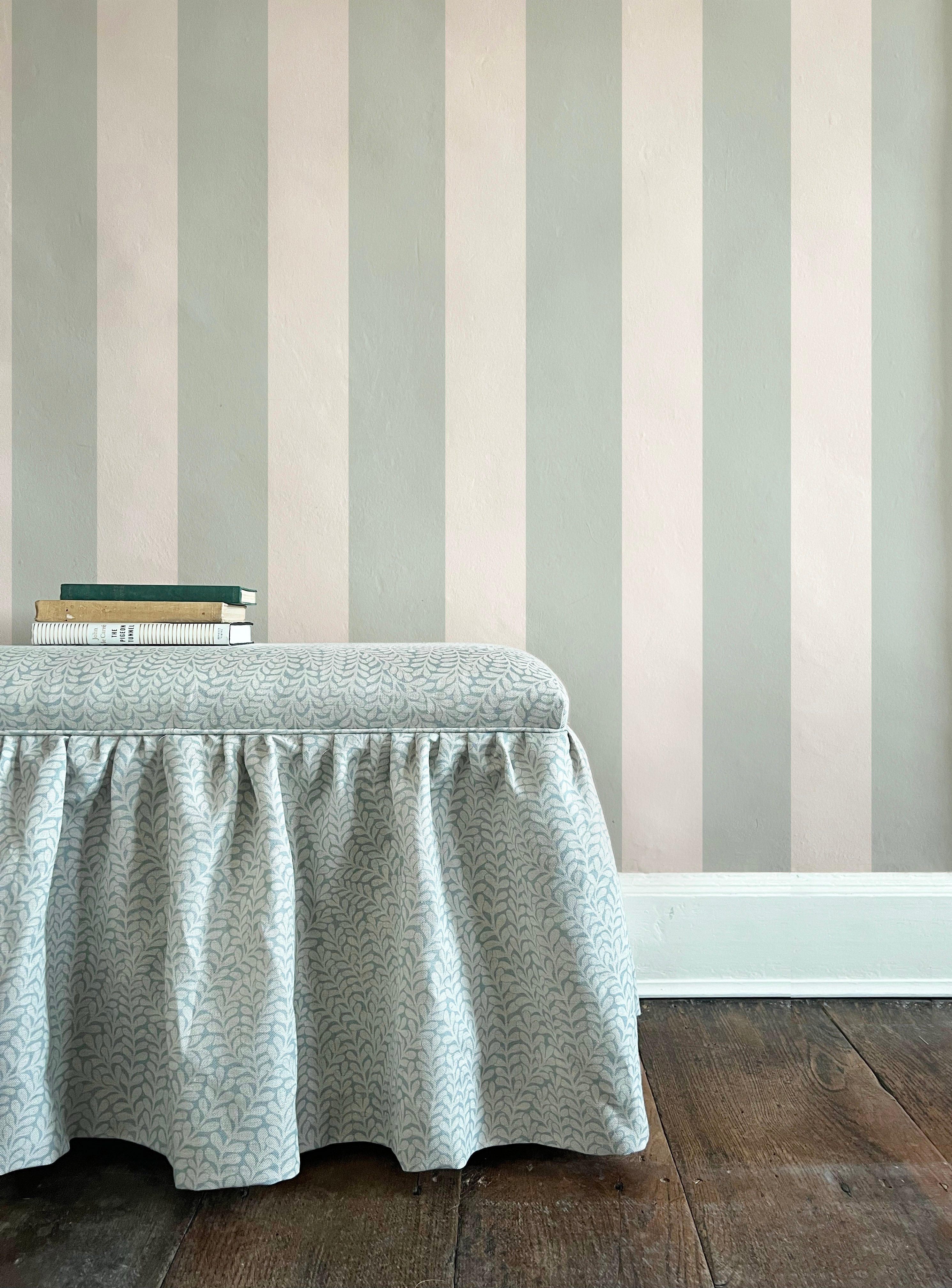 An upholstered ottoman topped with a stack of books stands in front of a wall papered in a wide stripe print.