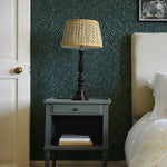 A cozy bedroom tableau with an end table, lamp and a statement wall papered in a scrolling stem and leaf print.