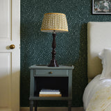 A cozy bedroom tableau with an end table, lamp and a statement wall papered in a scrolling stem and leaf print.