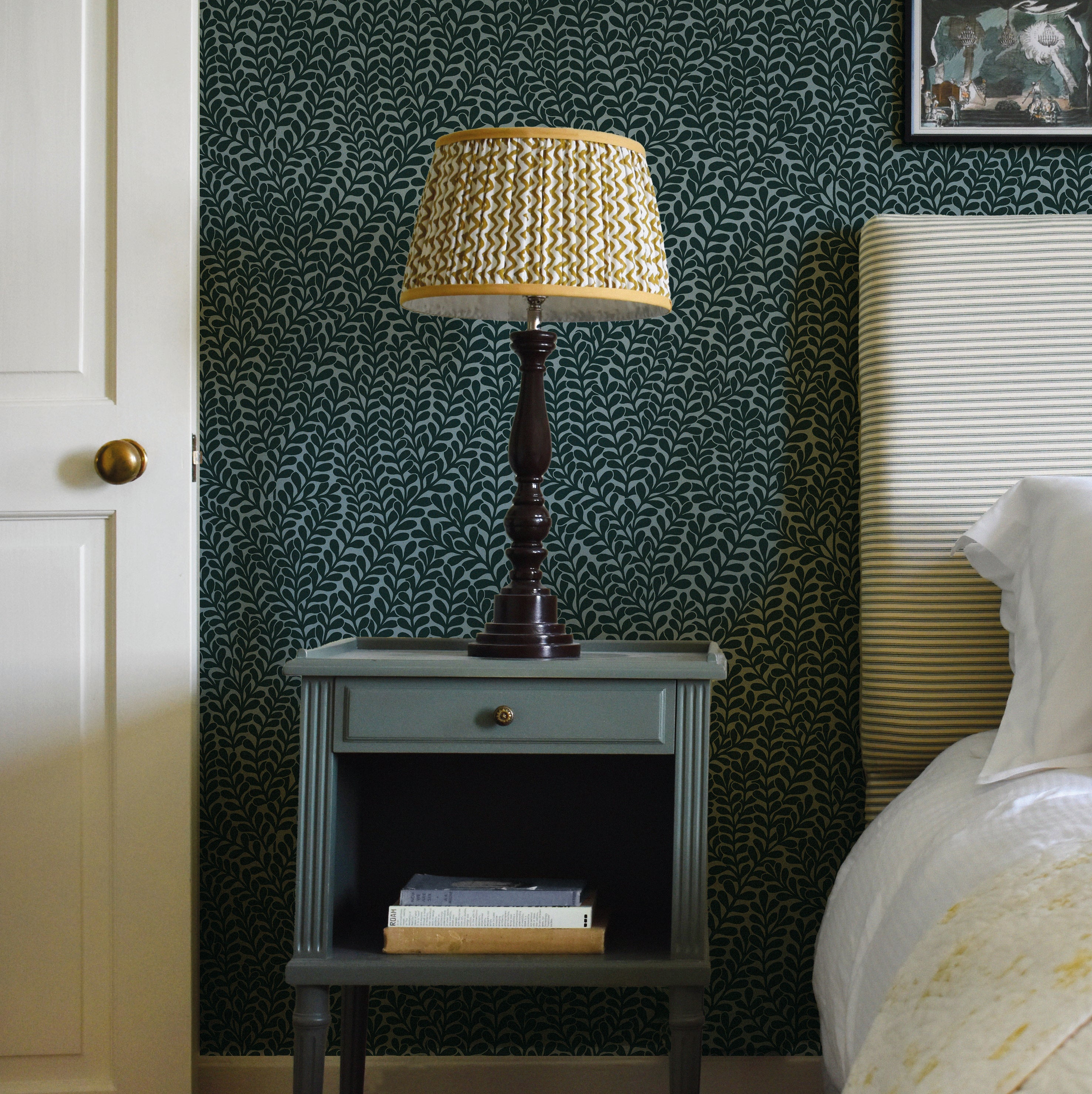 A cozy bedroom tableau with an end table, lamp and a statement wall papered in a scrolling stem and leaf print.