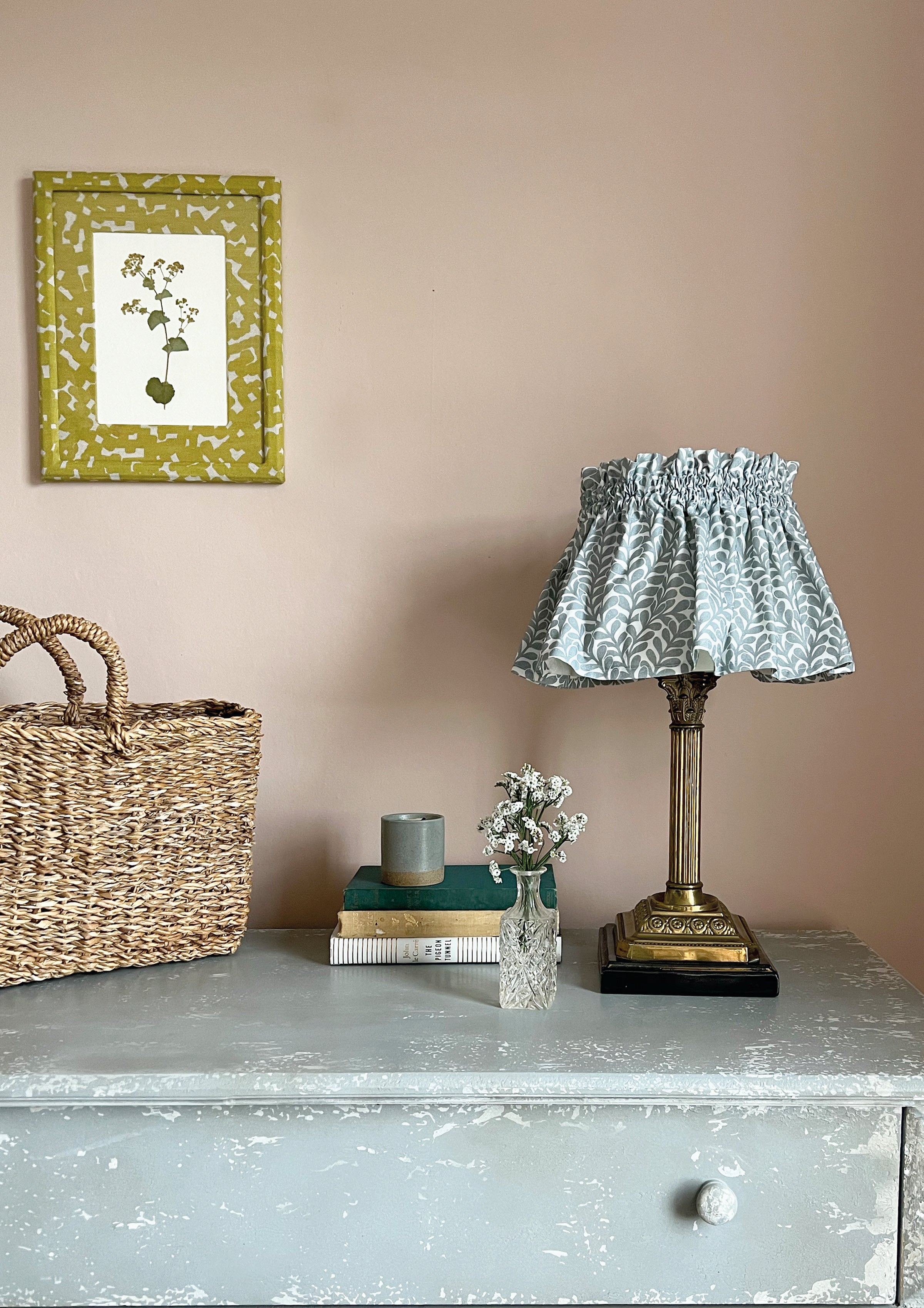 A dresser topped with a basket, books, and a lamp with a shade upholstered in a scrolling stem and leaf print.