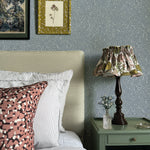 A cozy bedroom tableau with an end table, lamp and a statement wall papered in a scrolling stem and leaf print.
