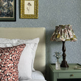A cozy bedroom tableau with an end table, lamp and a statement wall papered in a scrolling stem and leaf print.