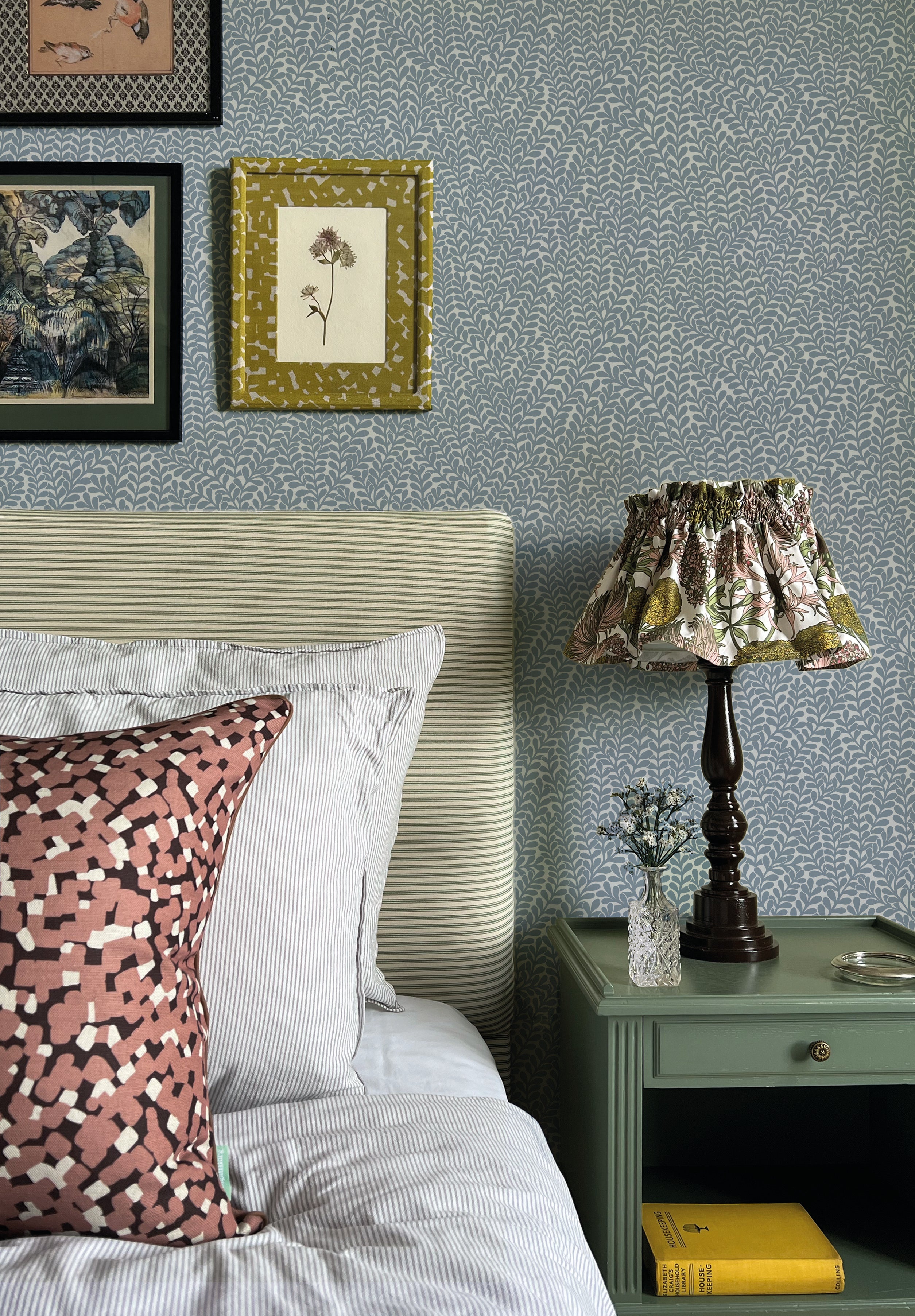 A cozy bedroom tableau with an end table, lamp and a statement wall papered in a scrolling stem and leaf print.