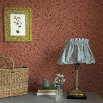 A maximalist end table with a basket, lamp, books and flowers in front of a wall papered in a scrolling stem and leaf print.