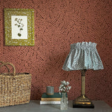 A maximalist end table with a basket, lamp, books and flowers in front of a wall papered in a scrolling stem and leaf print.