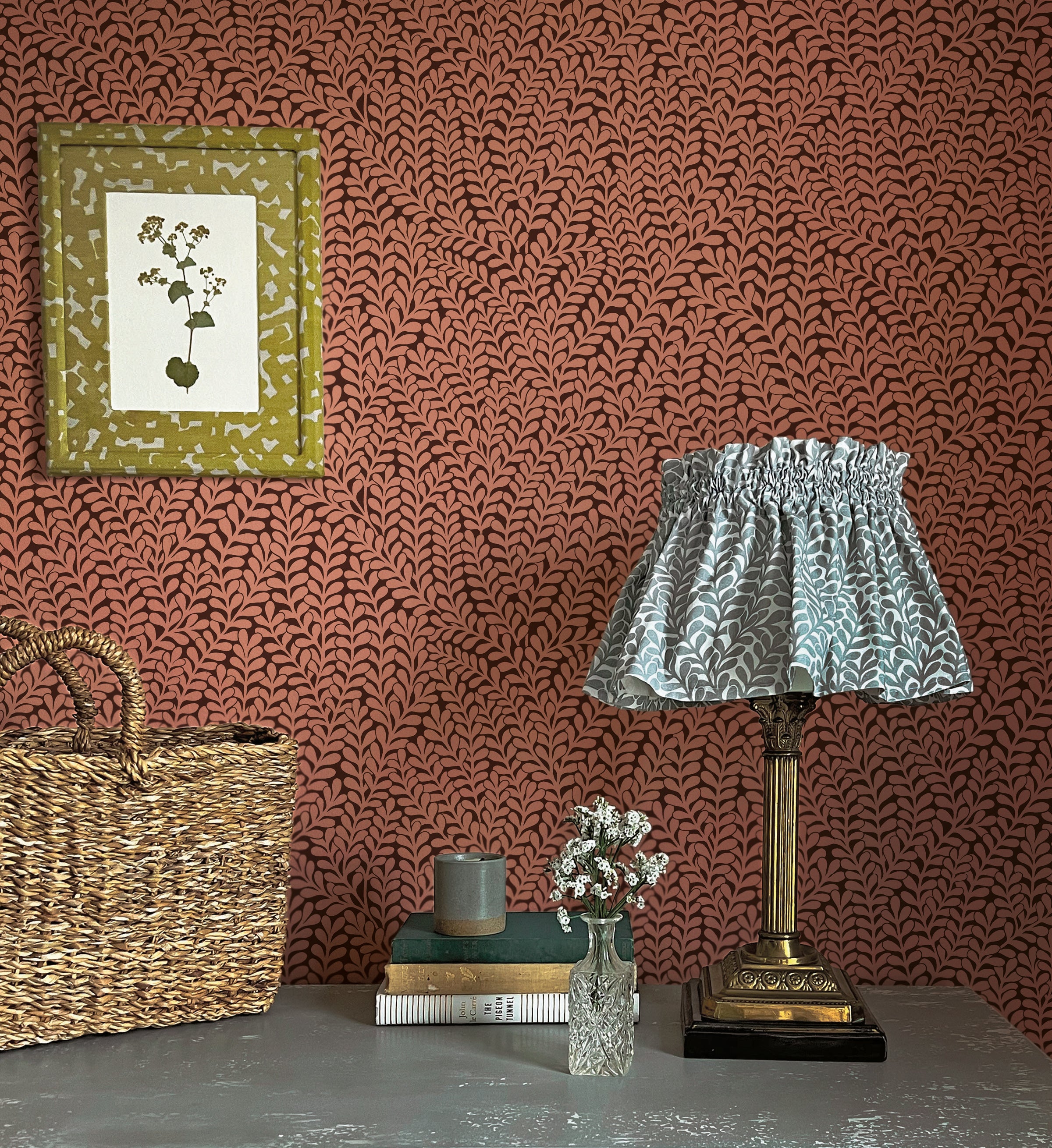 A maximalist end table with a basket, lamp, books and flowers in front of a wall papered in a scrolling stem and leaf print.
