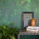 A large plant and an end table stand in front of a wall covered in multi-directional combed wallpaper in mottled green.