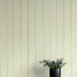 A vase of flowers stands in front of a wall covered in a playful irregular stripe print in light green on a cream field.
