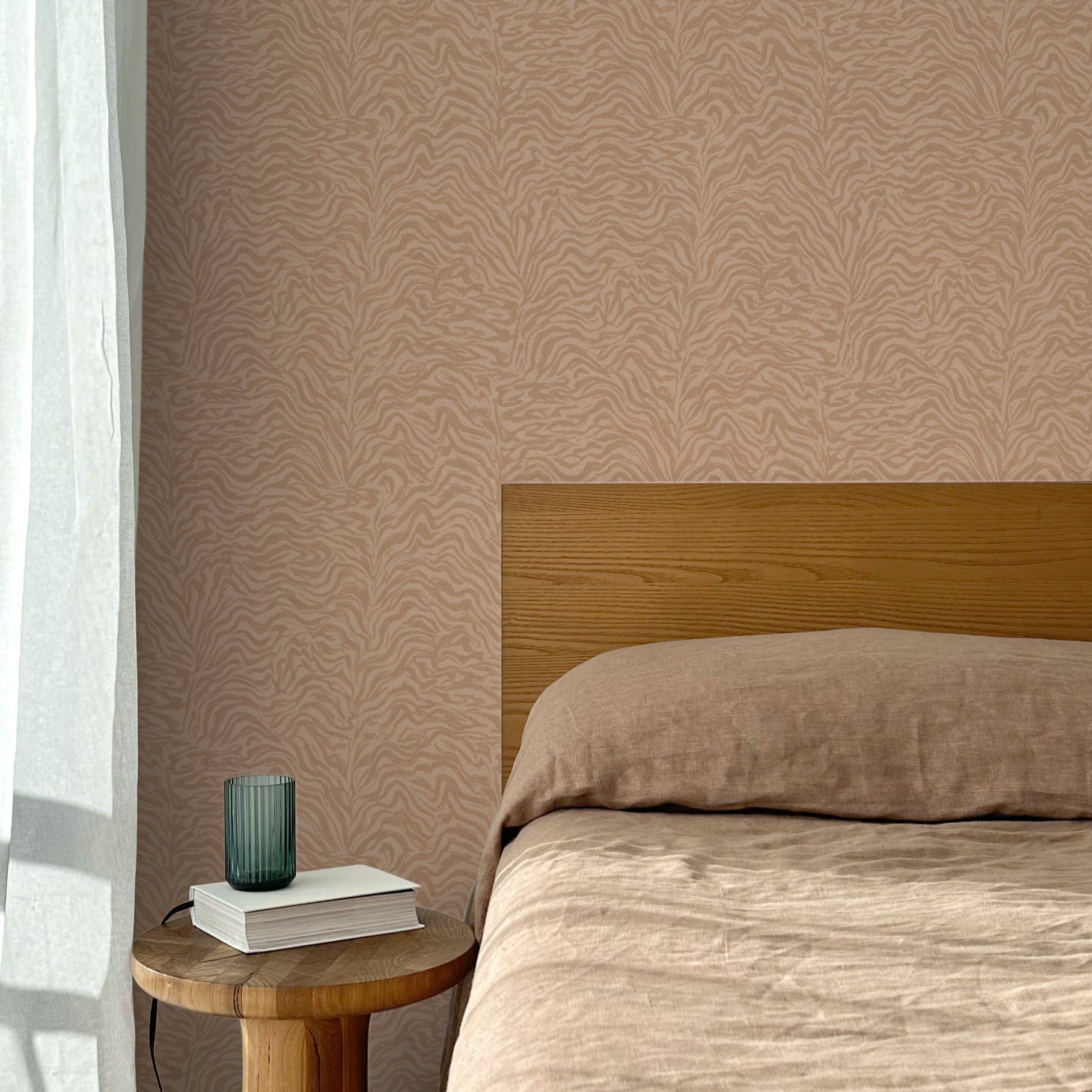 Bed with tan linens and wooden headboard, wooden side table  with white book and green glass, white floor length curtain against a watercolor swirling stripe wallpaper in tonal clay pink. 