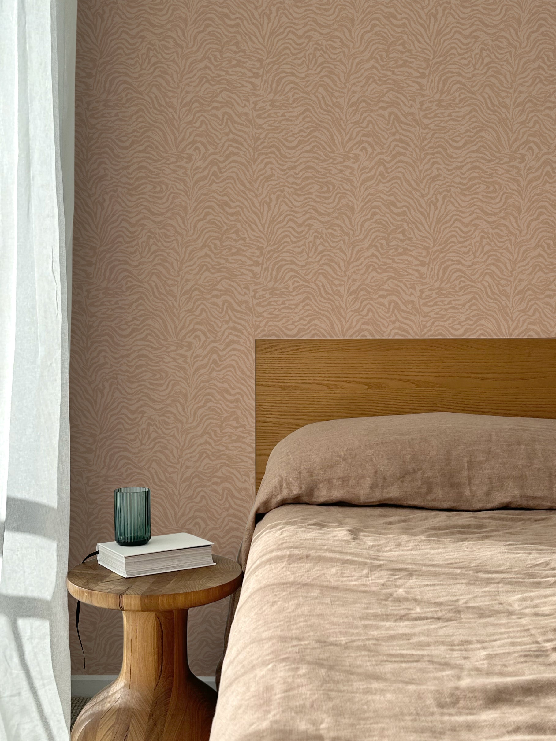 Bed with tan linens and wooden headboard, wooden side table  with white book and green glass, white floor length curtain against a watercolor swirling stripe wallpaper in tonal clay pink. 
