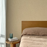 Bed with tan linens and wooden headboard, wooden side table  with white book and green glass, white floor length curtain against a watercolor swirling stripe wallpaper in tonal tan. 