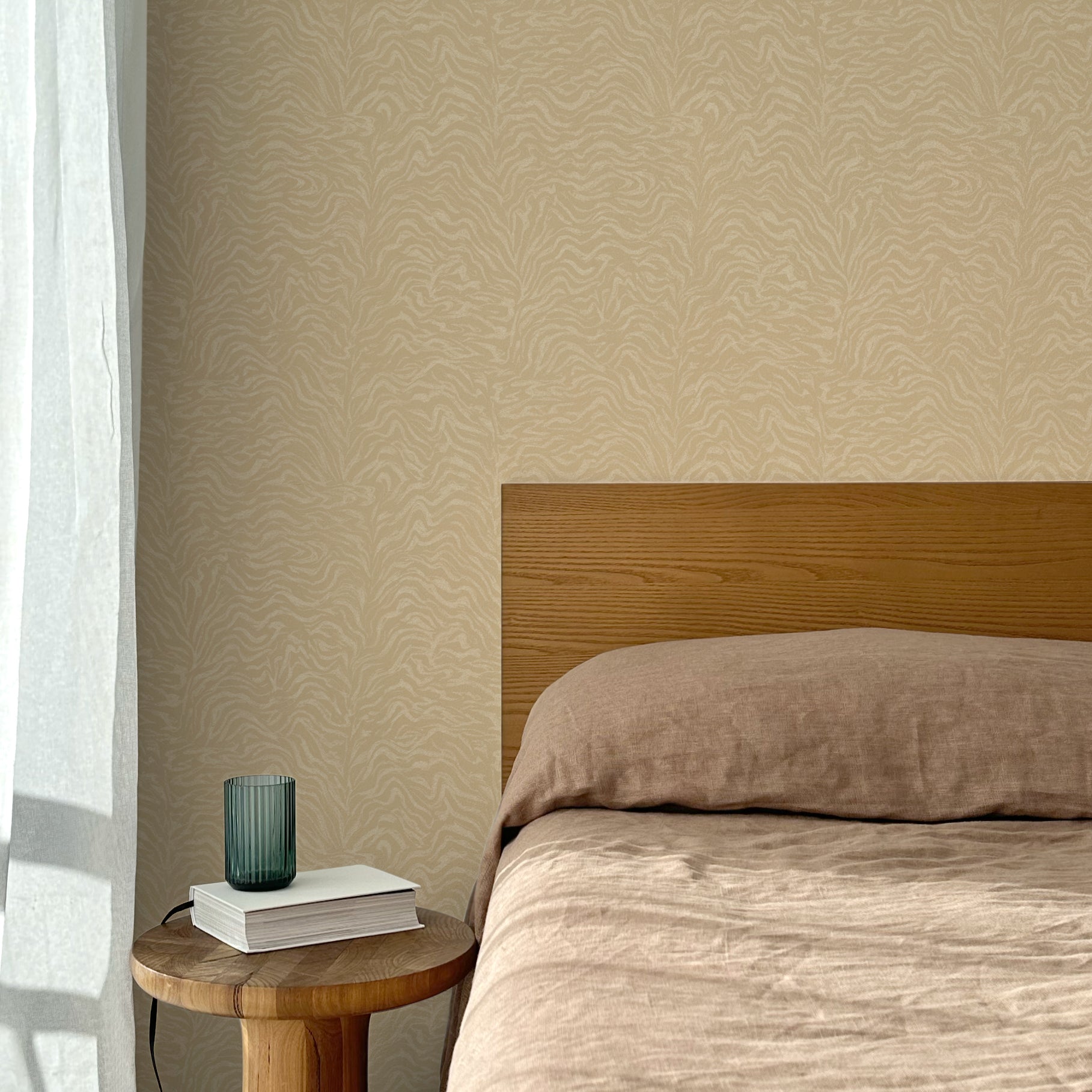 Bed with tan linens and wooden headboard, wooden side table  with white book and green glass, white floor length curtain against a watercolor swirling stripe wallpaper in tonal tan. 
