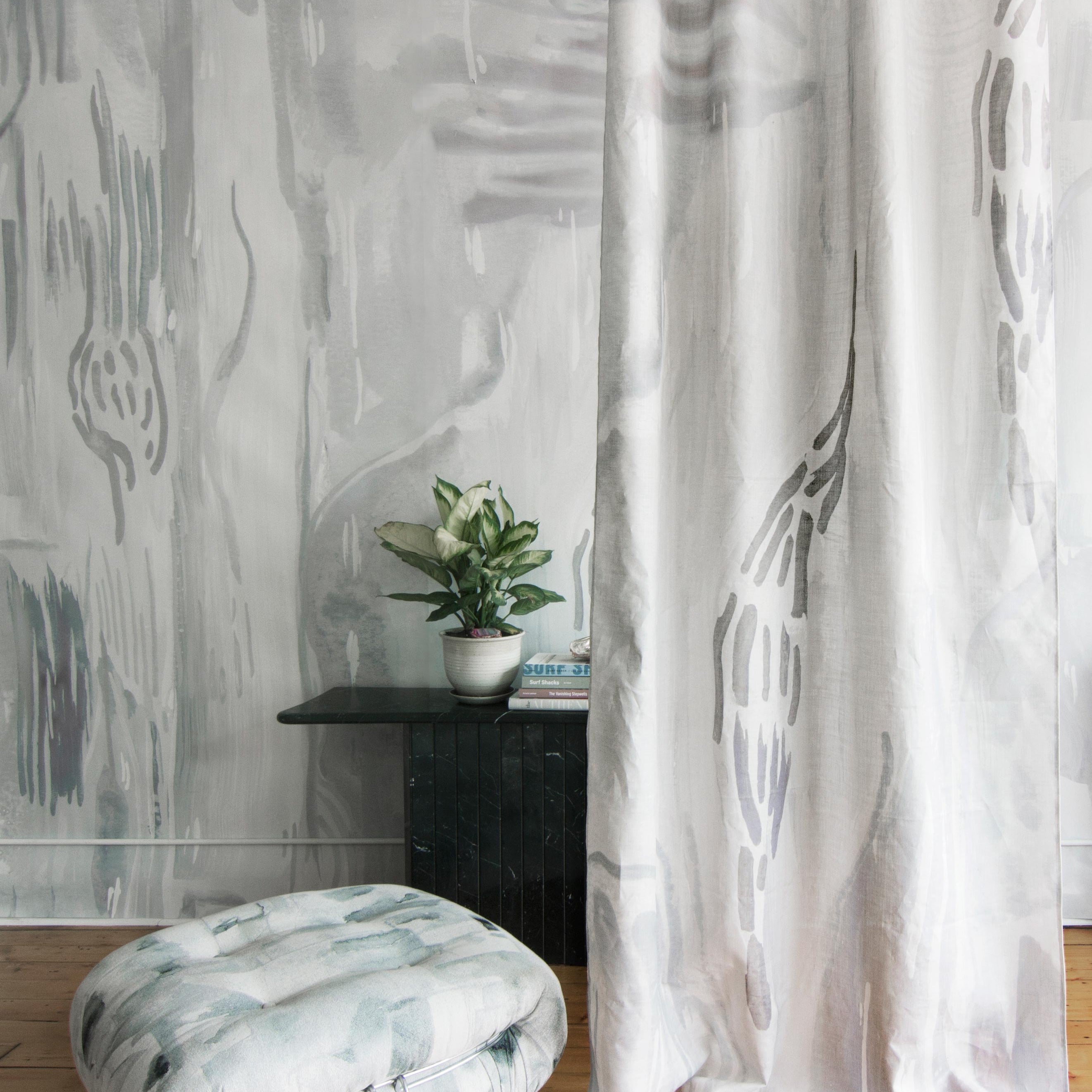 A floor pillow and curtain, both upholstered in coordinating watercolor paint prints in shades of gray and blue on white fields.