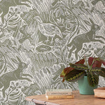 A wooden table with plant and books against a wall papered in a playful rabbit and bird print in white on a moss green field.