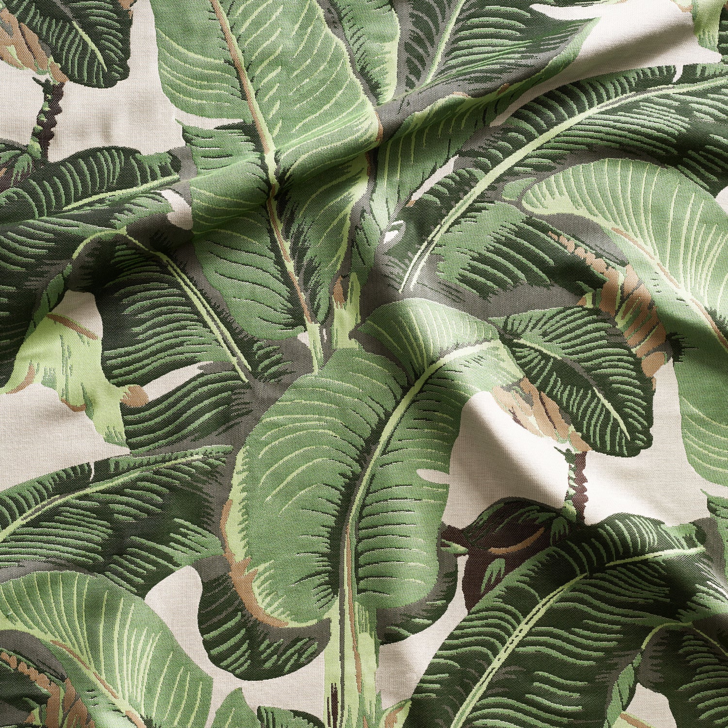 Close up of gently draped woven Indoor-Outdoor fabric with bold banana leaf in shades of green and tan on cream. 
