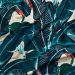 Close up of gently draped printed linen with bold banana leaf in shades of teal, navy and turquoise blue with green and coral accents on white.