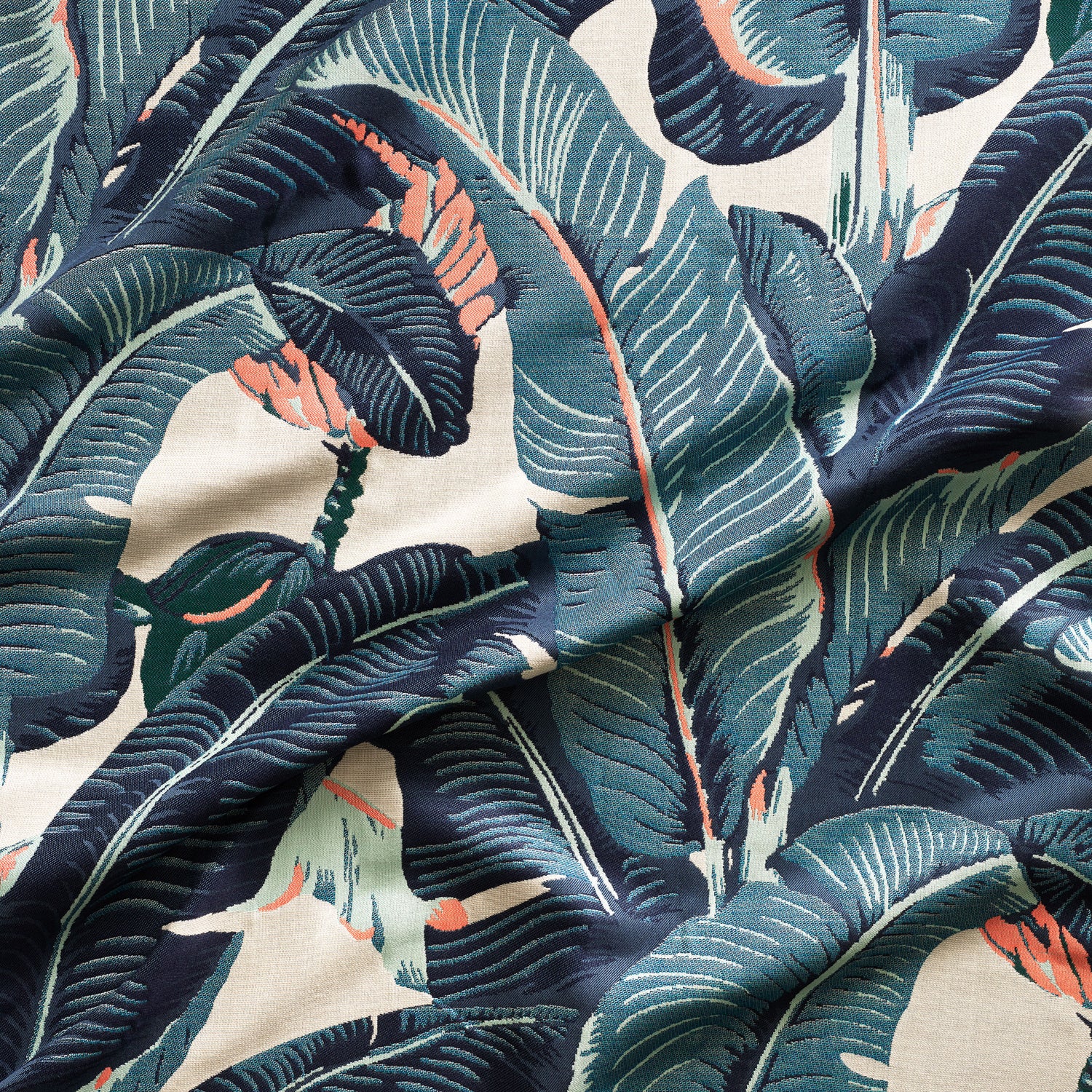 Close up of gently draped woven Indoor-Outdoor fabric with bold banana leaf in shades of teal blue and coral on beige.