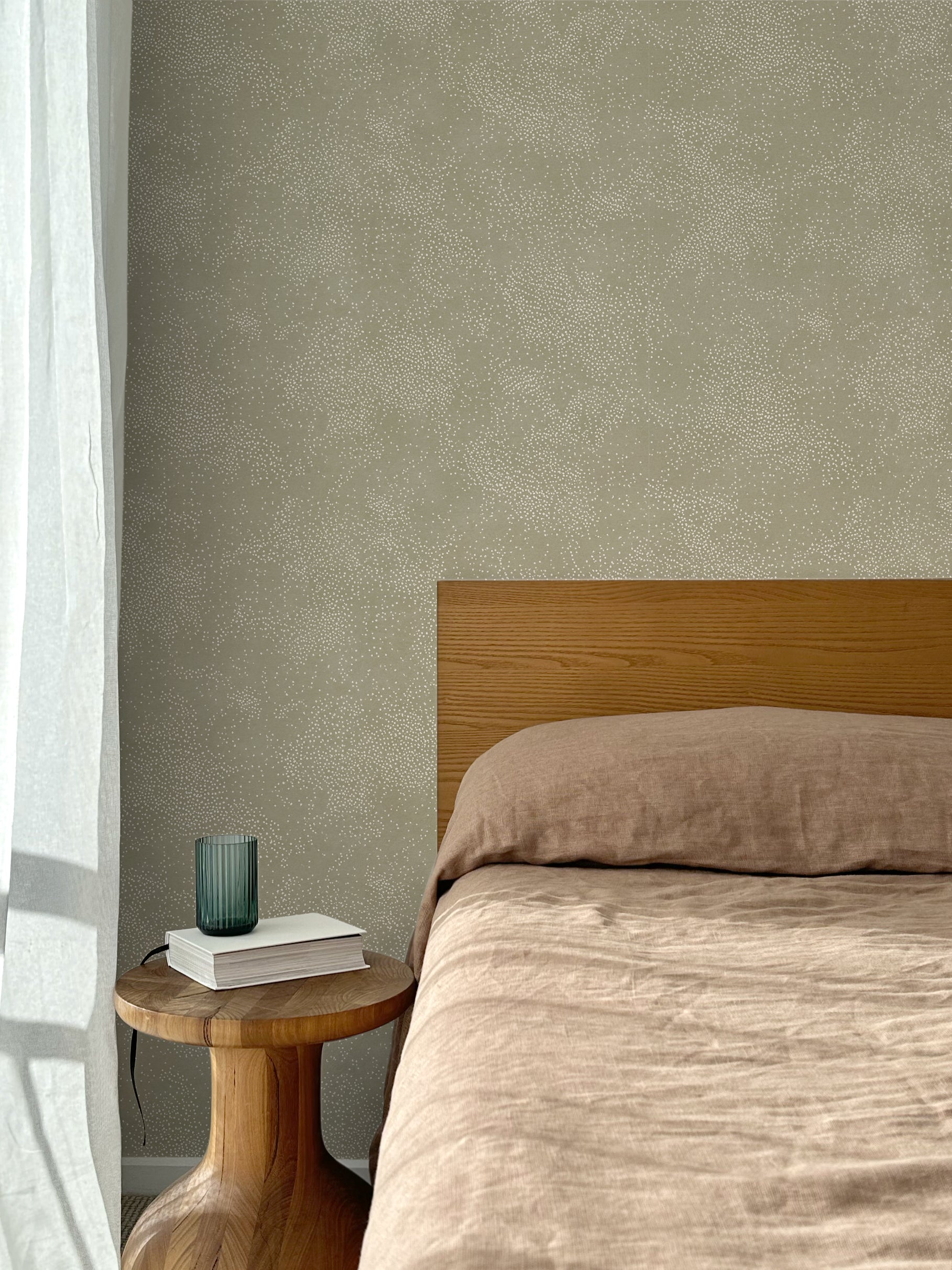 Bed with tan linens and wooden headboard, wooden side table  with white book and green glass, white floor length curtain against a white celestial dotted pattern on a sage green background. 