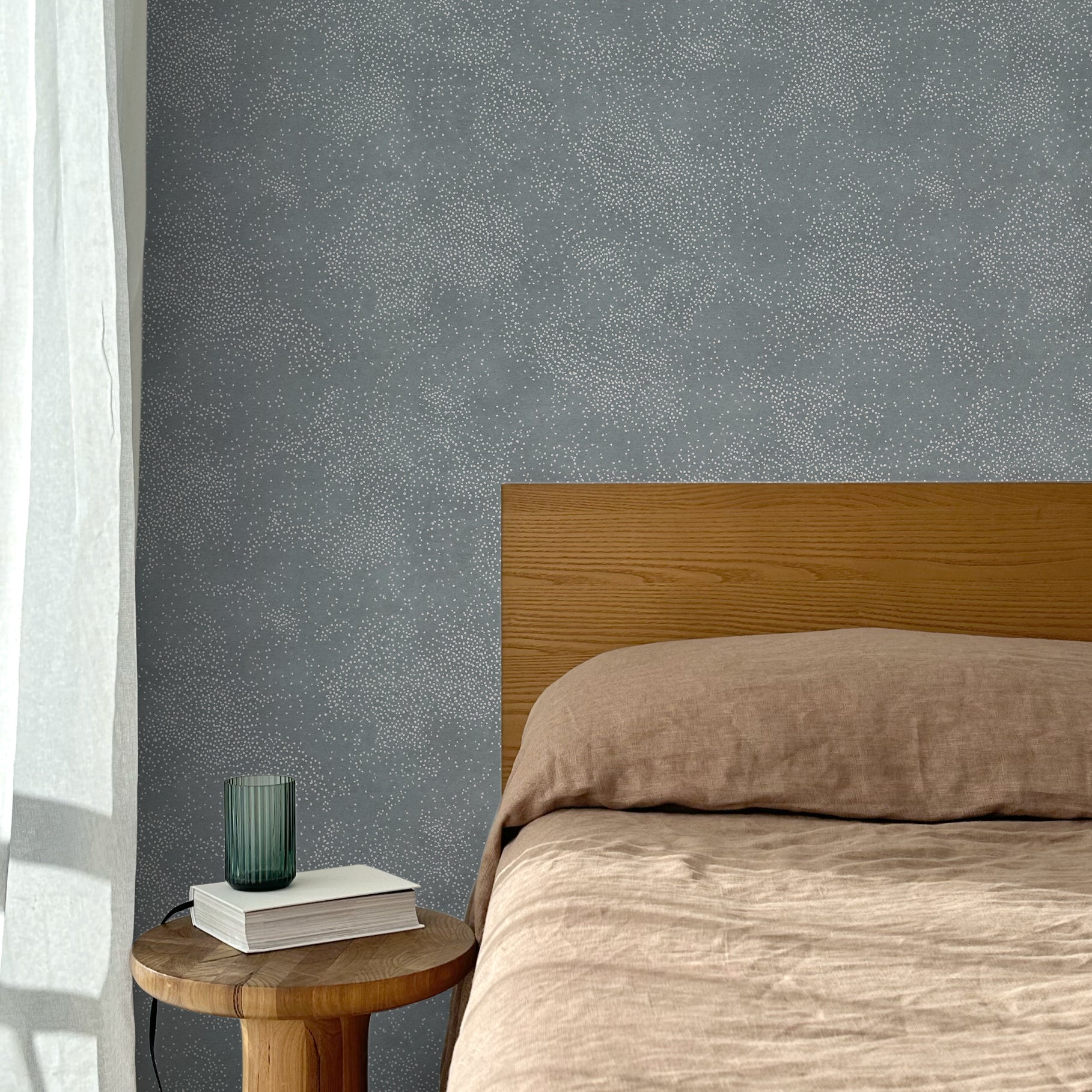 Bed with tan linens and wooden headboard, wooden side table  with white book and green glass, white floor length curtain against a white celestial dotted pattern on a cool blue background. 