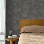 Bed with tan linens and wooden headboard, wooden side table  with white book and green glass, white floor length curtain against a white celestial dotted pattern on a charcoal grey background. 