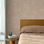 Bed with tan linens and wooden headboard, wooden side table  with white book and green glass, white floor length curtain against a white celestial dotted pattern on a clay pink background.