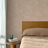 Bed with tan linens and wooden headboard, wooden side table  with white book and green glass, white floor length curtain against a white celestial dotted pattern on a clay pink background.