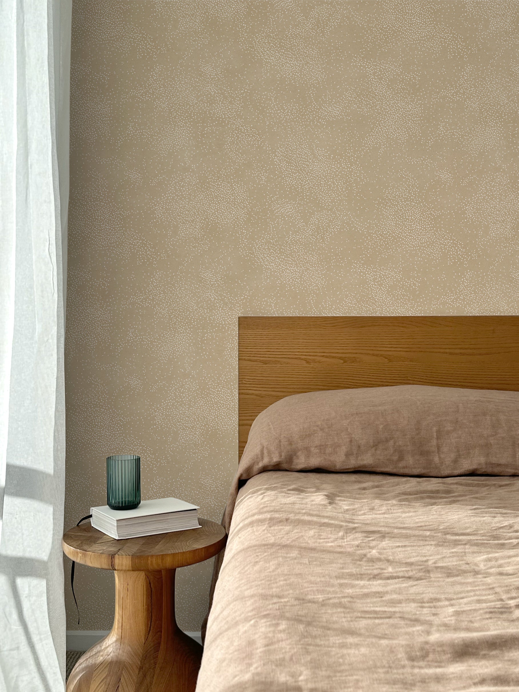 Bed with tan linens and wooden headboard, wooden side table  with white book and green glass, white floor length curtain against a white celestial dotted pattern on a tan background. 