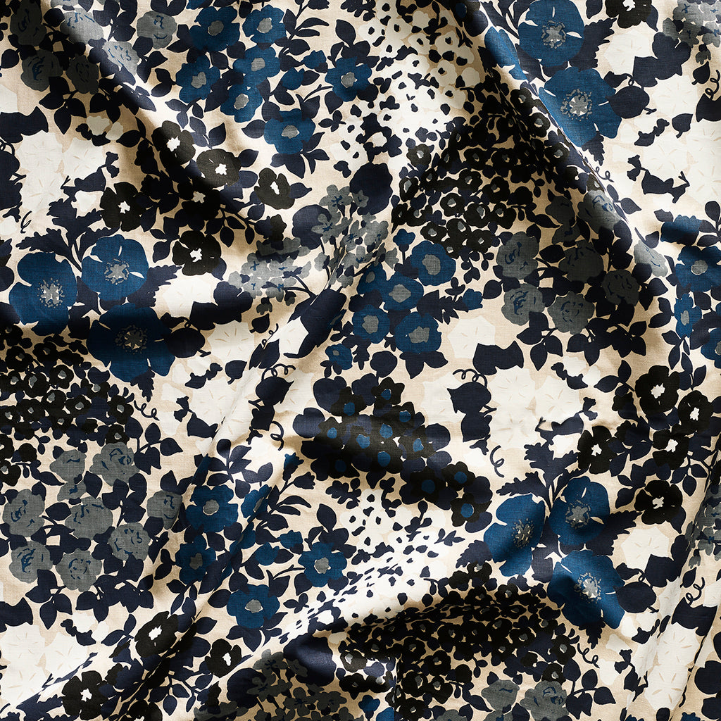 Close up of gently draped printed linen fabric with a dense flower and leaf pattern in shades of dark blue, grey, white and black on a beige background.