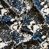 Close up of gently draped printed linen fabric with a dense flower and leaf pattern in shades of dark blue, grey, white and black on a beige background.
