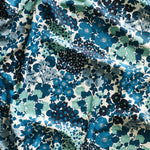 Close up of gently draped printed linen fabric with a dense flower and leaf pattern in shades of blue and turquoise on a white background.