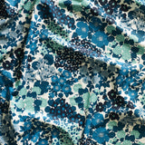Close up of gently draped printed linen fabric with a dense flower and leaf pattern in shades of blue and turquoise on a white background.