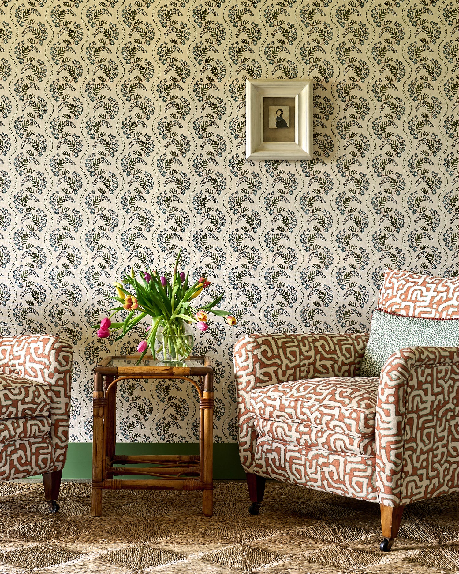 Living room scheme with orange and cream patterned chairs, sisal rug, rattan side table and a floral stripe wallpaper. 